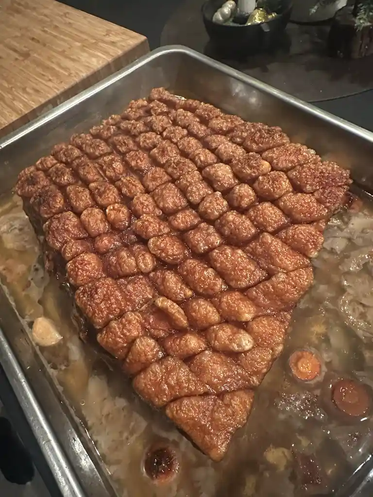 A close-up photo shows a large, roasted piece of meat in a metal baking tray, its surface scored into a grid of small…