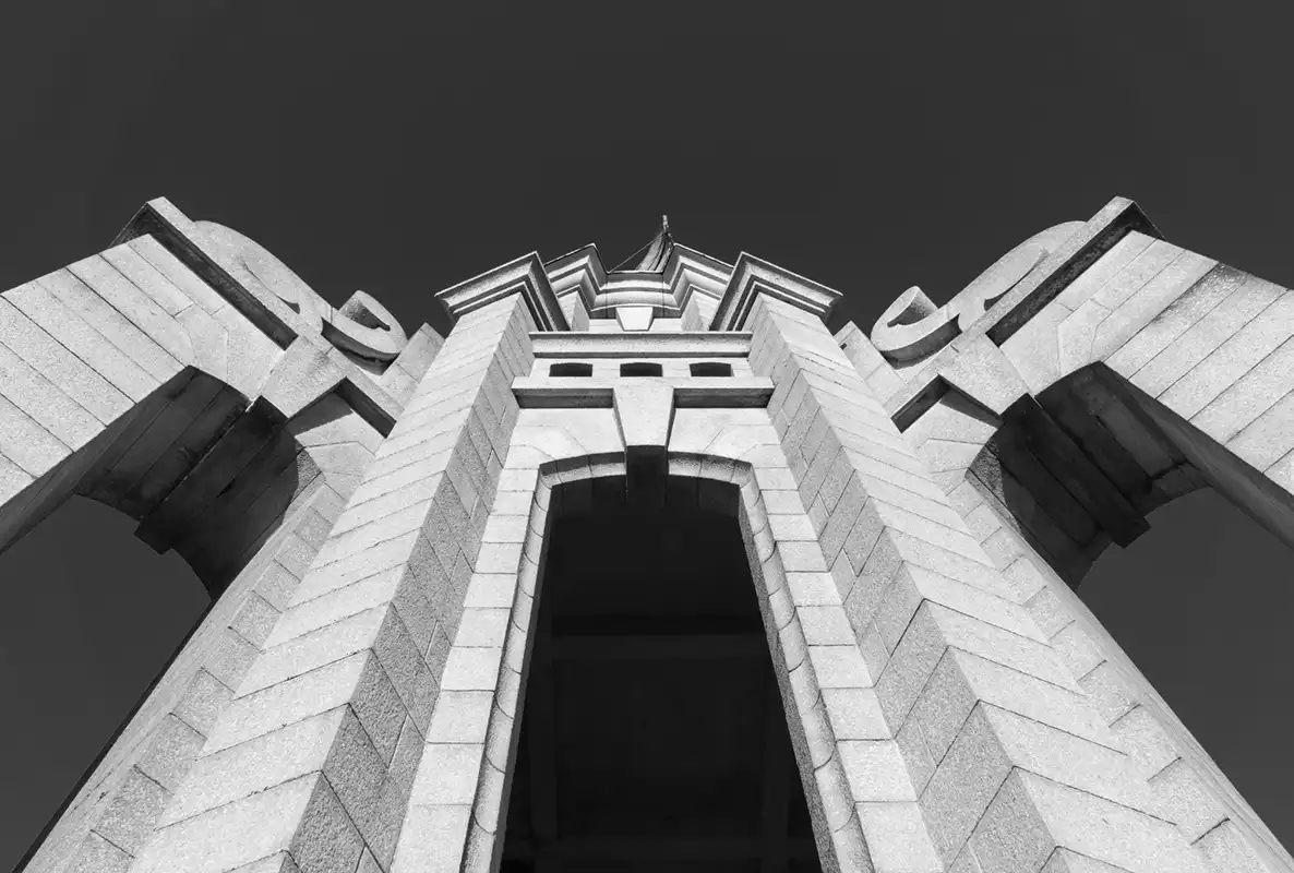 A black-and-white, low-angle photograph shows the imposing, symmetrical stone facade of a monumental building, possibly a…