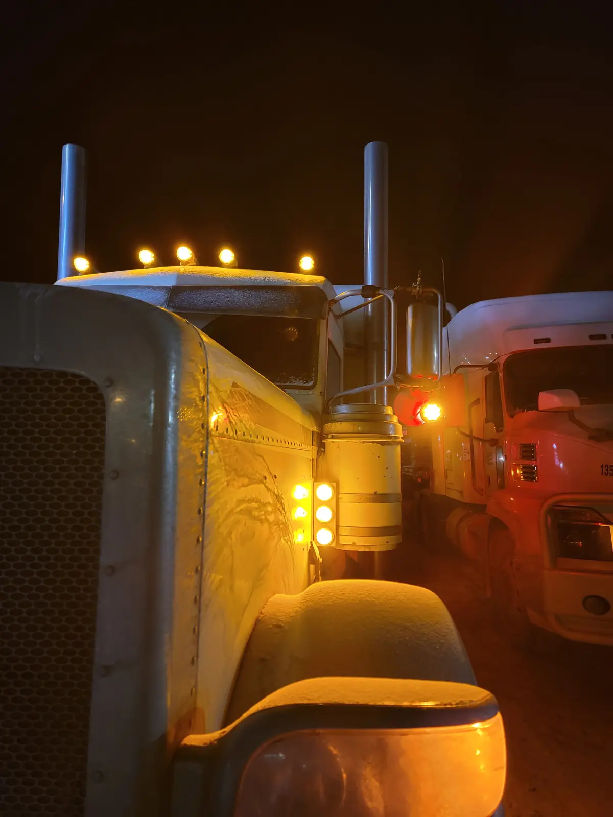 A close-up nighttime photo shows two large semi-trucks parked side-by-side, their surfaces covered in snow and ice. The…