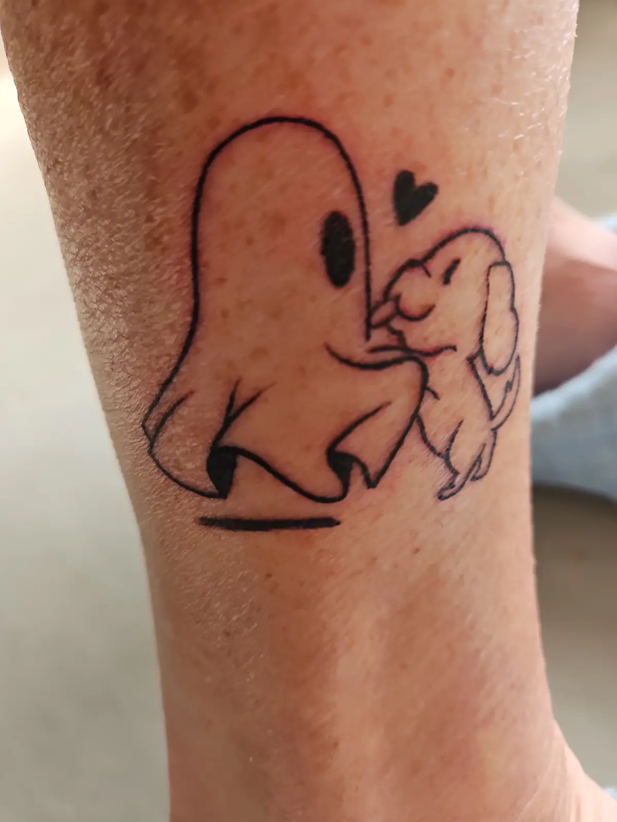 A black ink tattoo on a person's forearm depicts a simple cartoon ghost with two black eyes and a smiling mouth, leaning in…