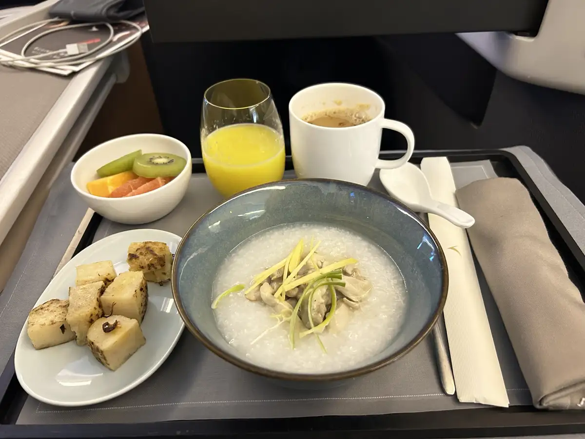 A close-up photo of a luxurious airplane breakfast tray featuring a bowl of congee, a plate of grilled cubes, a bowl of…