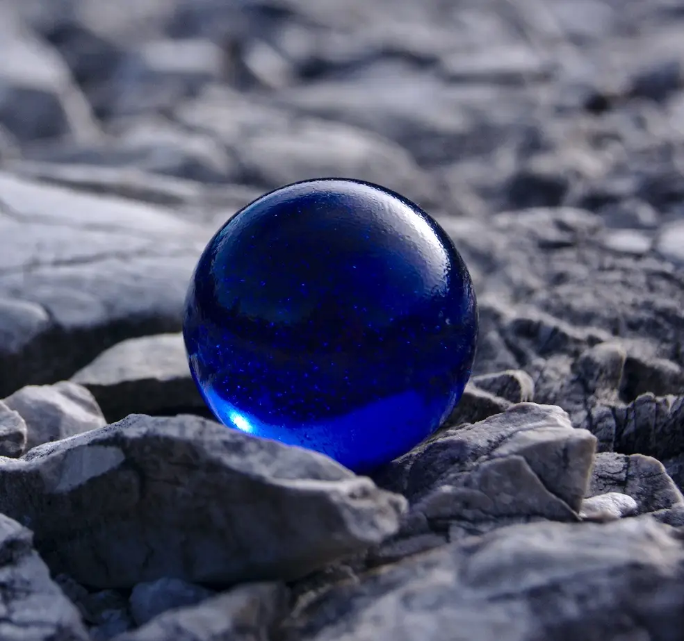 A glossy, deep blue glass marble with glittery specks sits nestled among gray, rough rocks. The marble reflects light and…