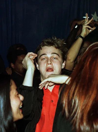 A young man with messy hair and a red shirt looks overwhelmed and uncomfortable at a crowded party, surrounded by people…