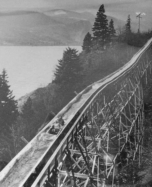A black-and-white historical photo shows two people riding a wooden roller coaster track that descends steeply over a…