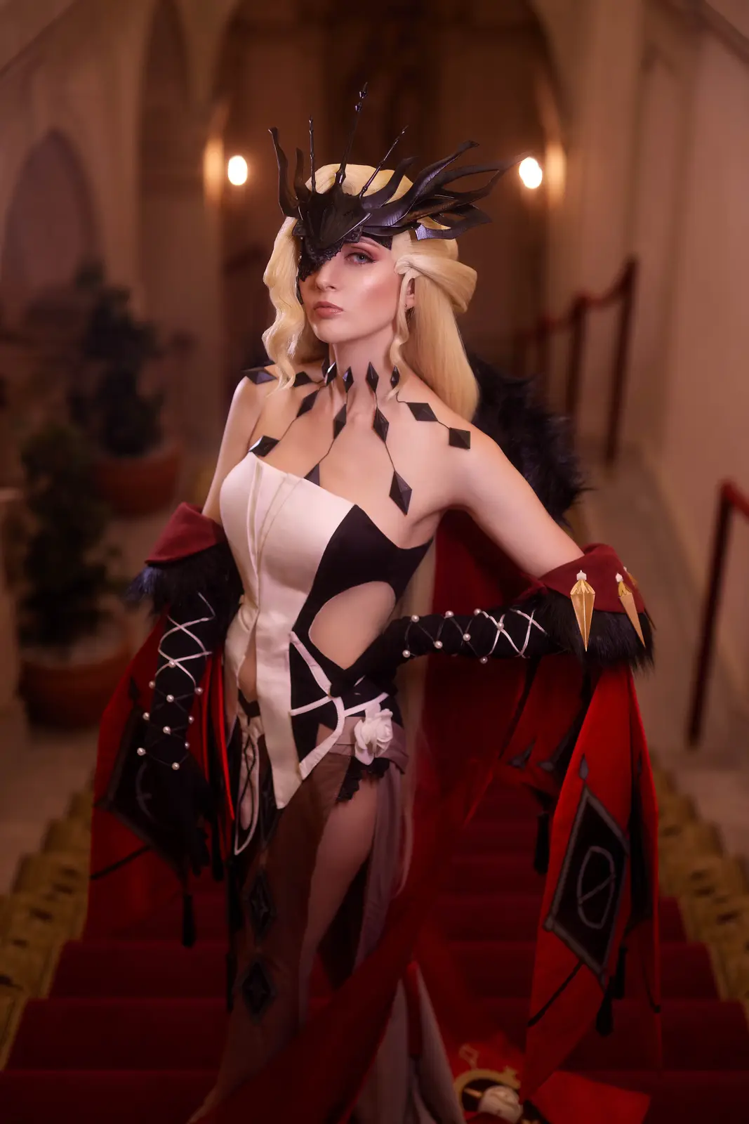 A cosplayer in an elaborate black, white, and red costume with a spiked crown and eye mask stands confidently on a…