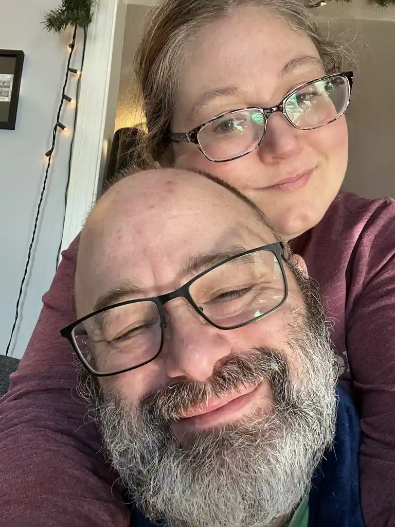A selfie shows a bald man with a gray beard and black-rimmed glasses smiling with his eyes closed, while a woman with…