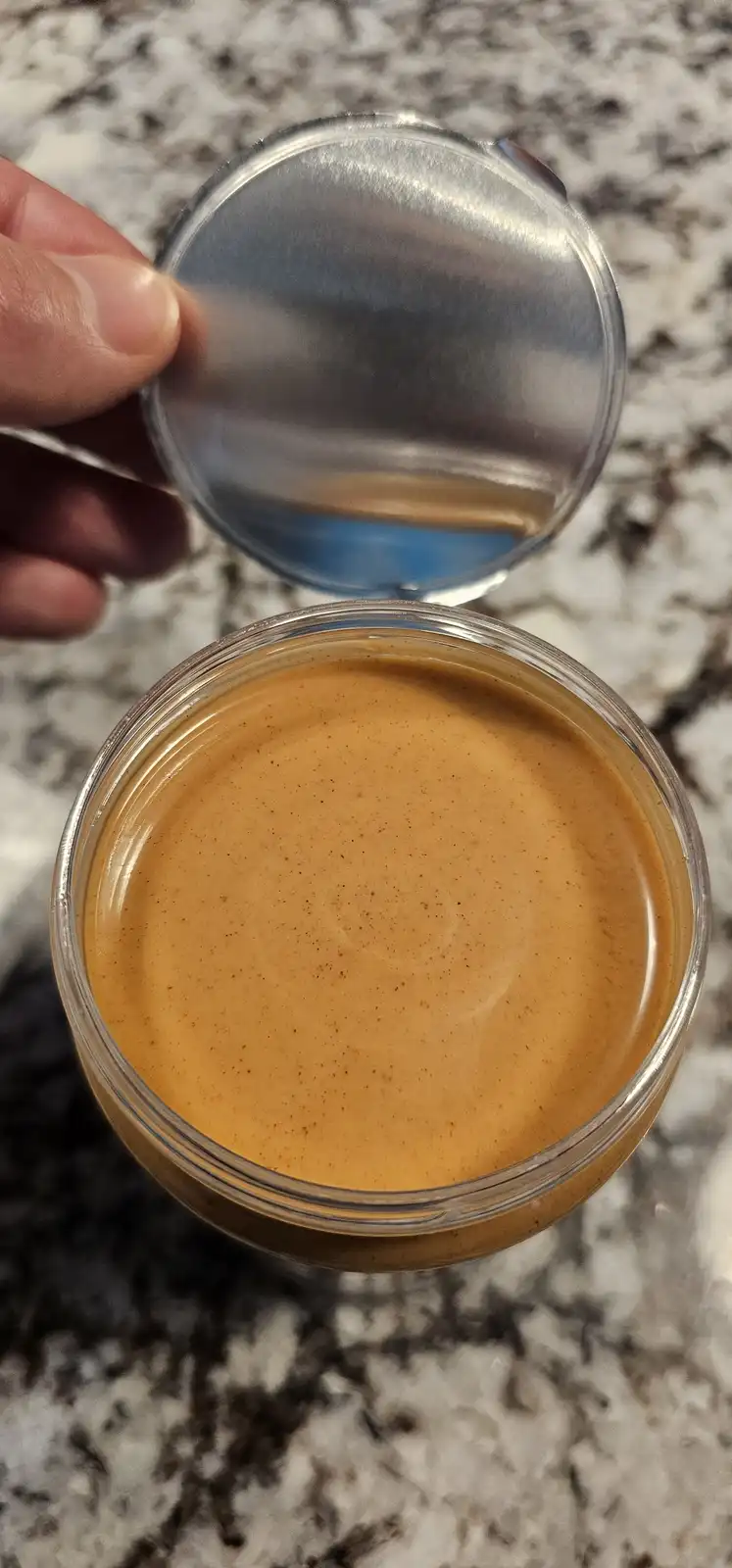 A hand holds the lid off a clear glass jar filled with a smooth, light brown liquid resembling coffee creamer, set on a…