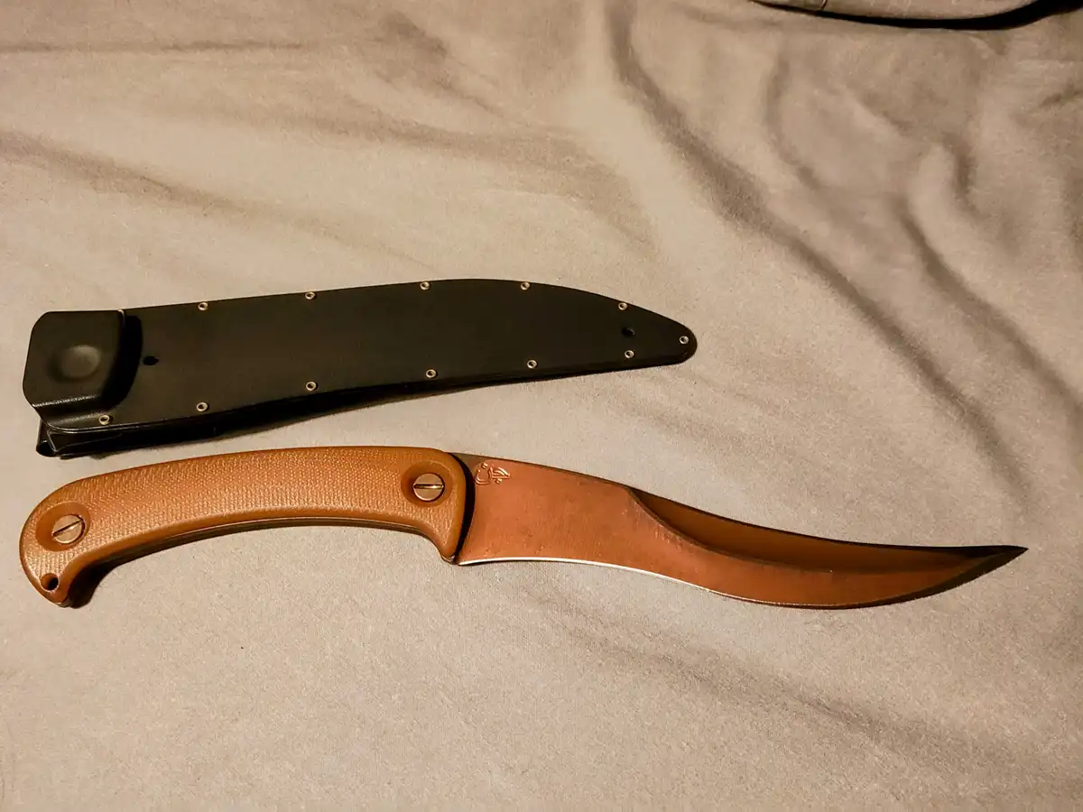 A copper-colored fixed-blade knife with a textured handle lies next to its black sheath on a light gray fabric surface. The…