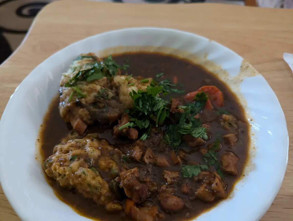 A close-up photo shows a white bowl filled with a dark brown stew containing chunks of meat, carrots, and beans, topped with…