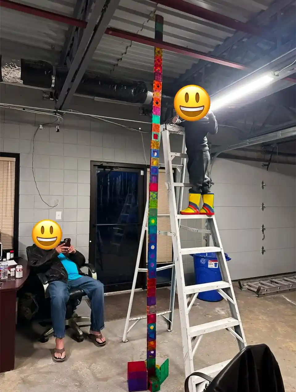 In a garage, one person sits in a chair holding a phone while another stands on a tall ladder, wearing bright yellow rain…