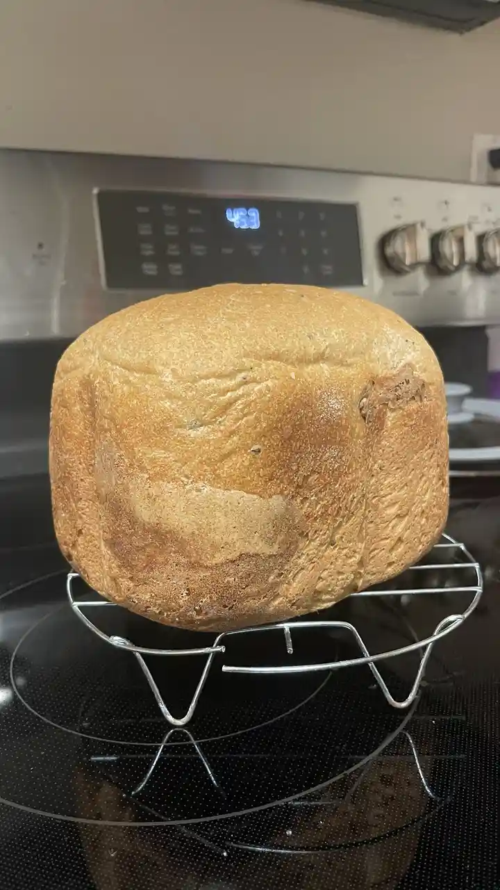 A round loaf of golden-brown bread sits on a metal cooling rack atop a black stovetop, with a digital oven display in the…