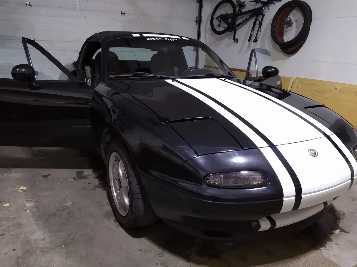 A black Mazda Miata convertible with white racing stripes is parked inside a garage, its driver's side door open. On the…