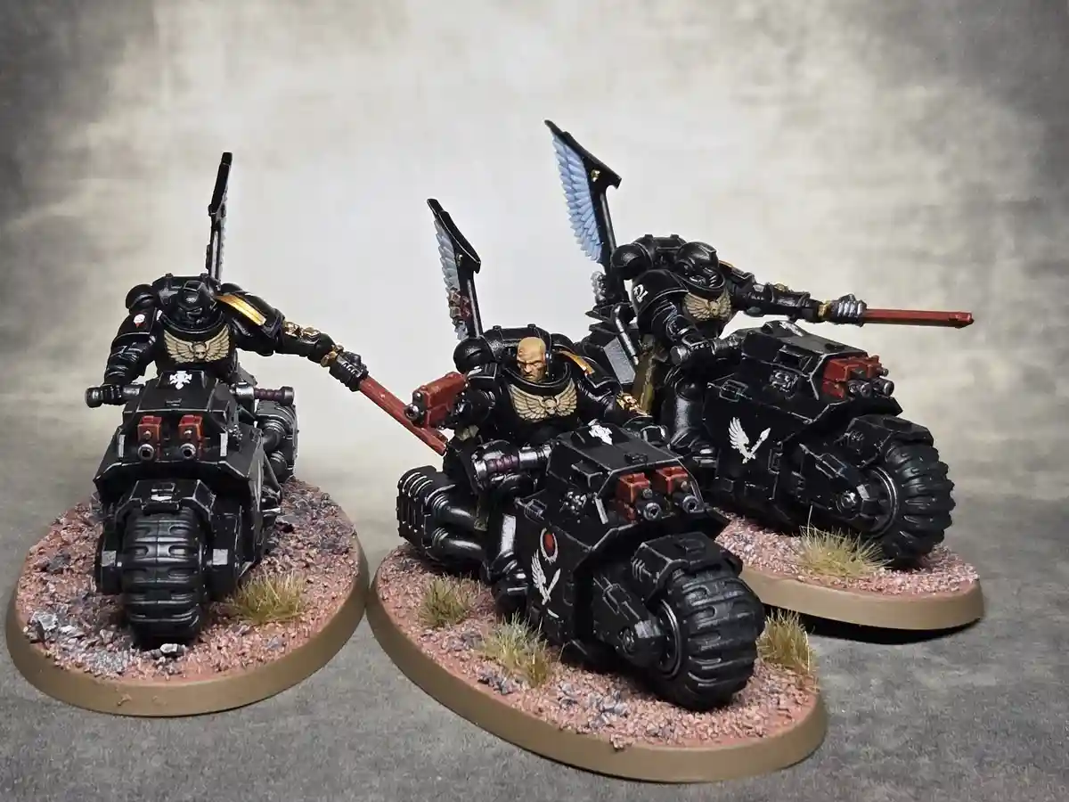 Three painted miniature figures of Space Marines on heavily armored motorcycles, each mounted on a circular base with gravel…