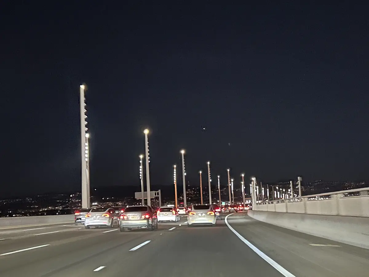 A nighttime photo from a car on a multi-lane highway, showing traffic ahead with brake lights glowing red. Tall, modern…