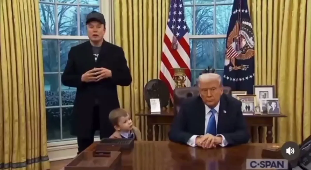 A man in a black coat and cap stands behind a desk in the Oval Office, gesturing with his hands as if speaking. Seated at…