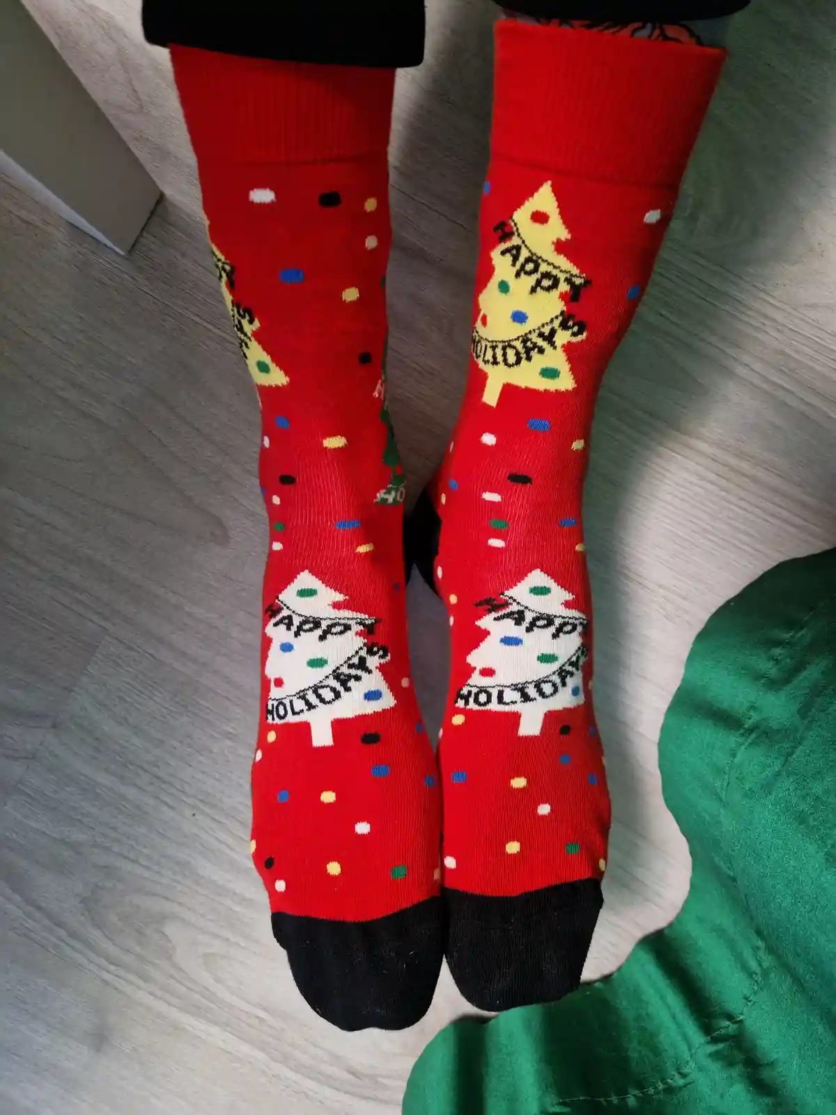 A pair of red holiday socks with black toes and cuffs, featuring colorful polka dots and Christmas trees with 'happy…
