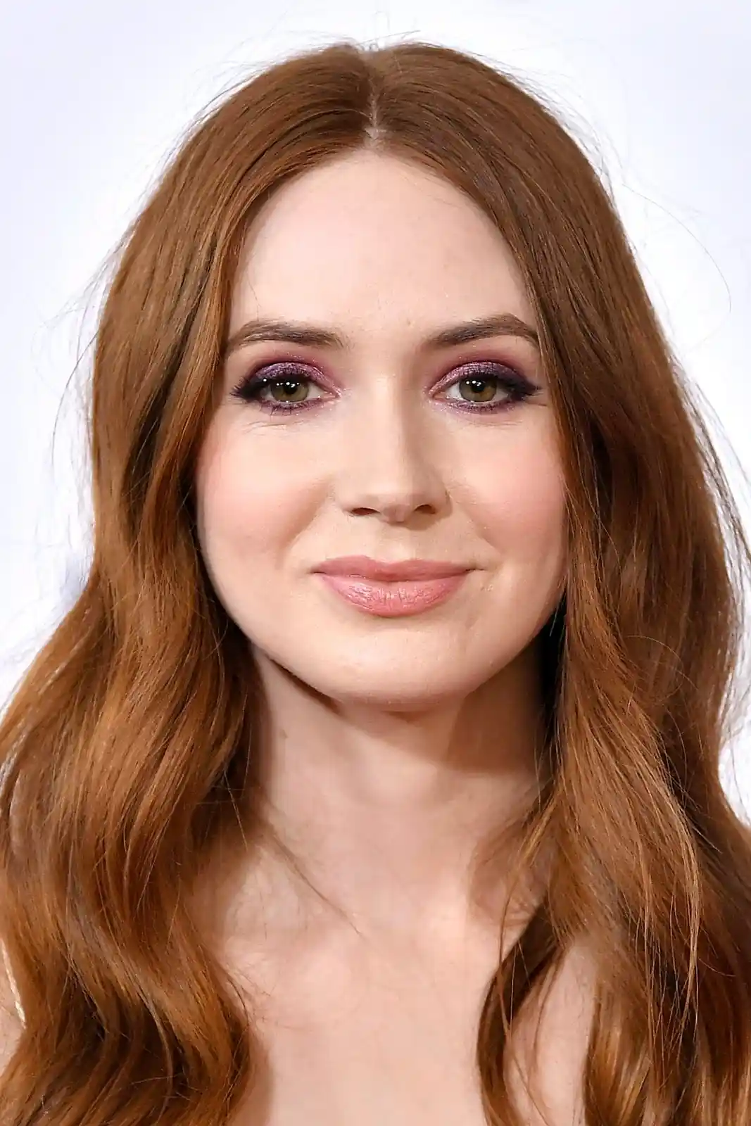 A close-up photo of a woman with long, wavy auburn hair and prominent purple eyeshadow, smiling slightly. The overlaid text…