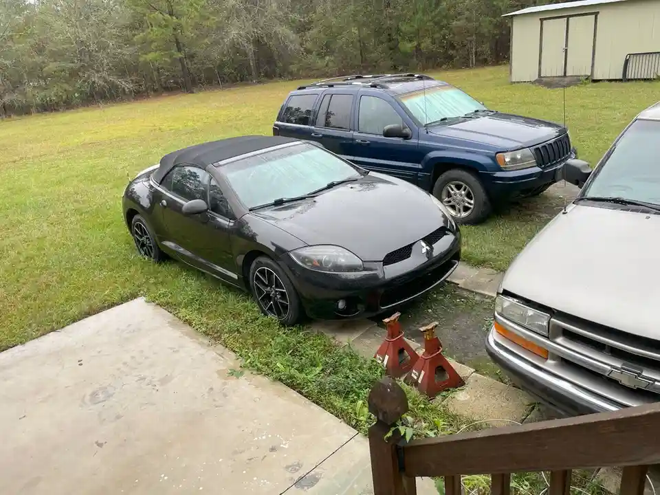 A black Mitsubishi Eclipse convertible is parked on grass next to a driveway, with two red jack stands placed on the…