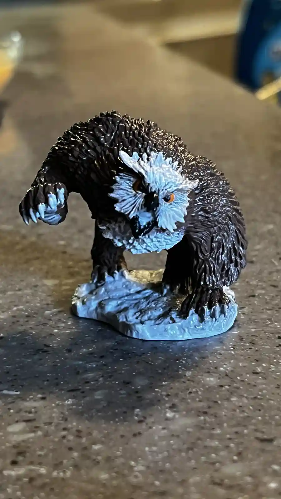 A small figurine of a bear-like creature with a distinctly owl-shaped head, featuring large orange eyes and feather-like…