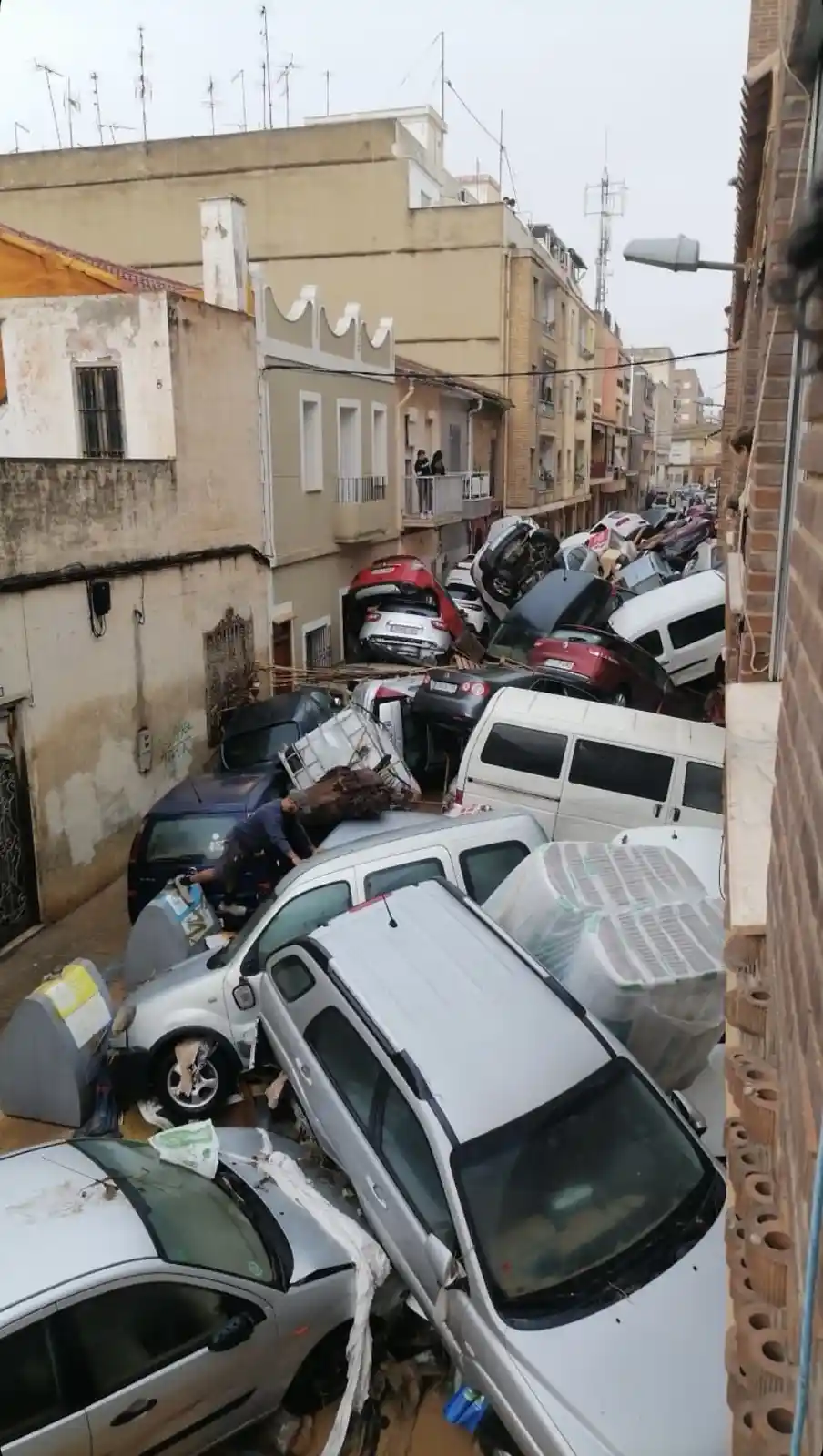 A narrow urban street is completely blocked by a chaotic pile of crushed, overturned, and stacked cars, with debris and…
