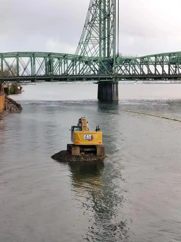 A yellow Caterpillar excavator sits partially submerged in water, resting on a small pile of rocks, with a large green truss…