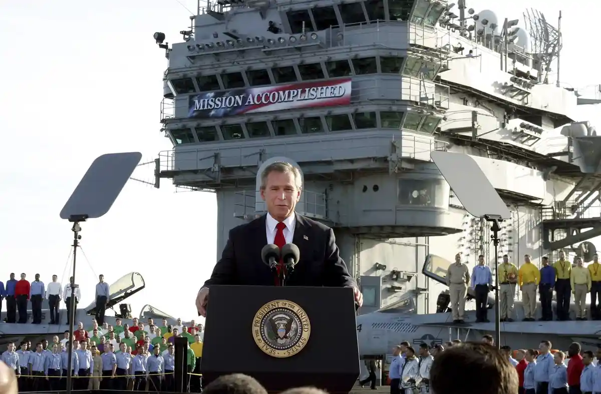 A man in a suit and red tie stands at a podium with the presidential seal, addressing a crowd on an aircraft carrier. Behind…