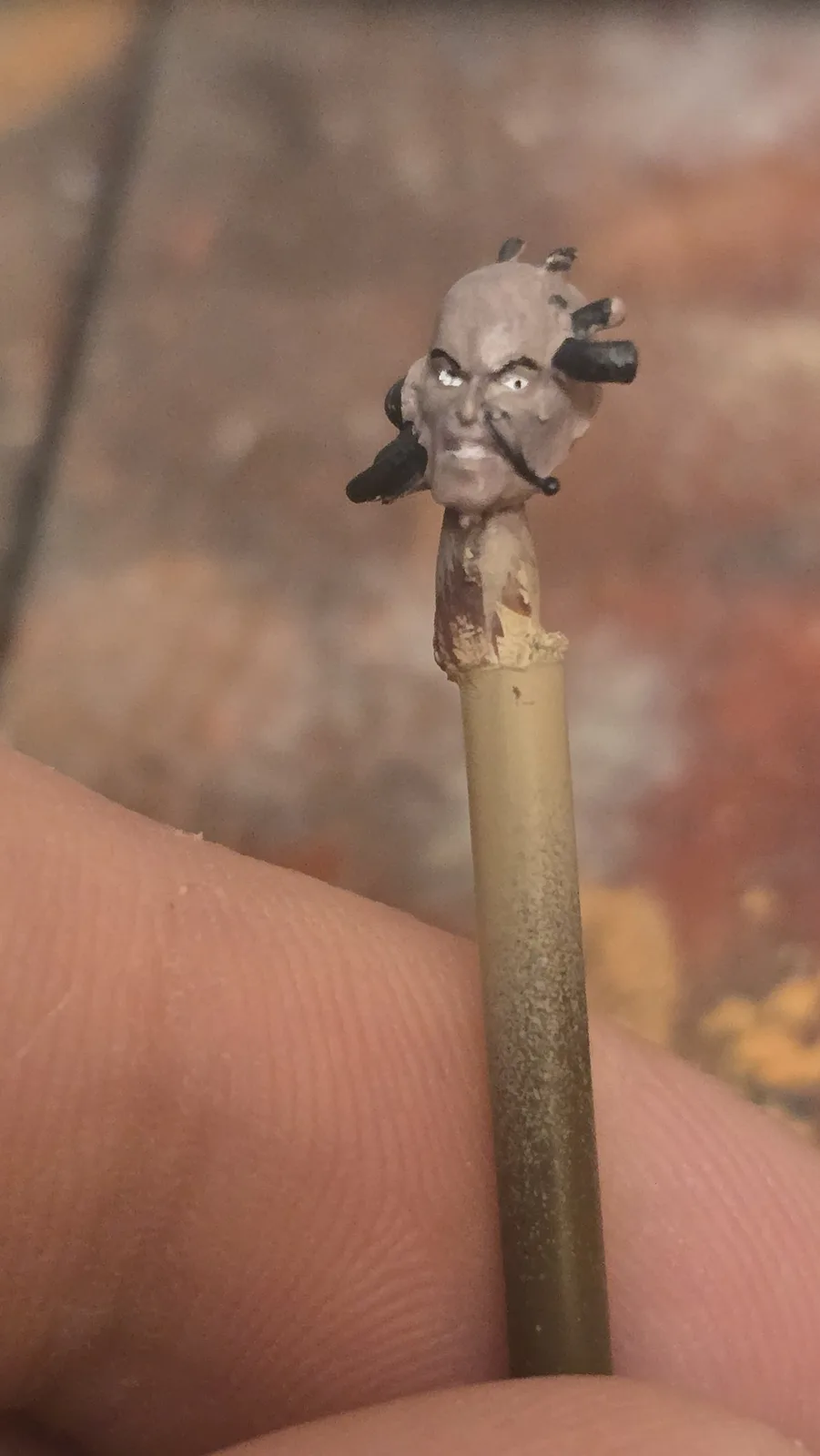 A close-up photo shows a person's fingers holding a pencil. At the top of the pencil is a small, crudely sculpted figurine…
