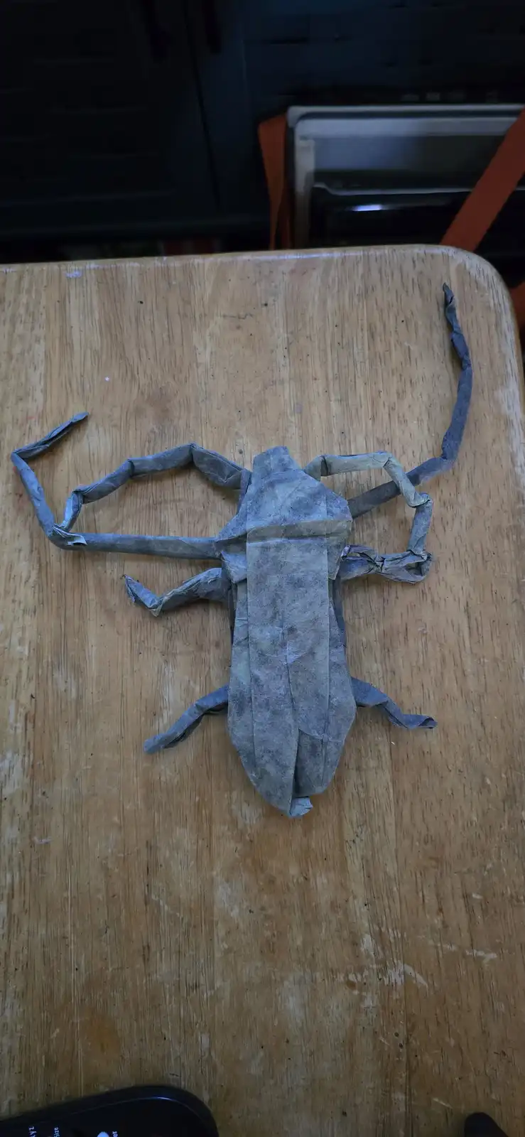 A gray origami scorpion with long, segmented legs and a curved tail rests on a worn wooden table. The overlaid text reads 'i…