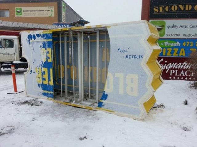 A large, broken, snow-covered sign for 'BLOK' (likely 'BLOCKBUSTER') lies on its side in a snowy parking lot, with the text…