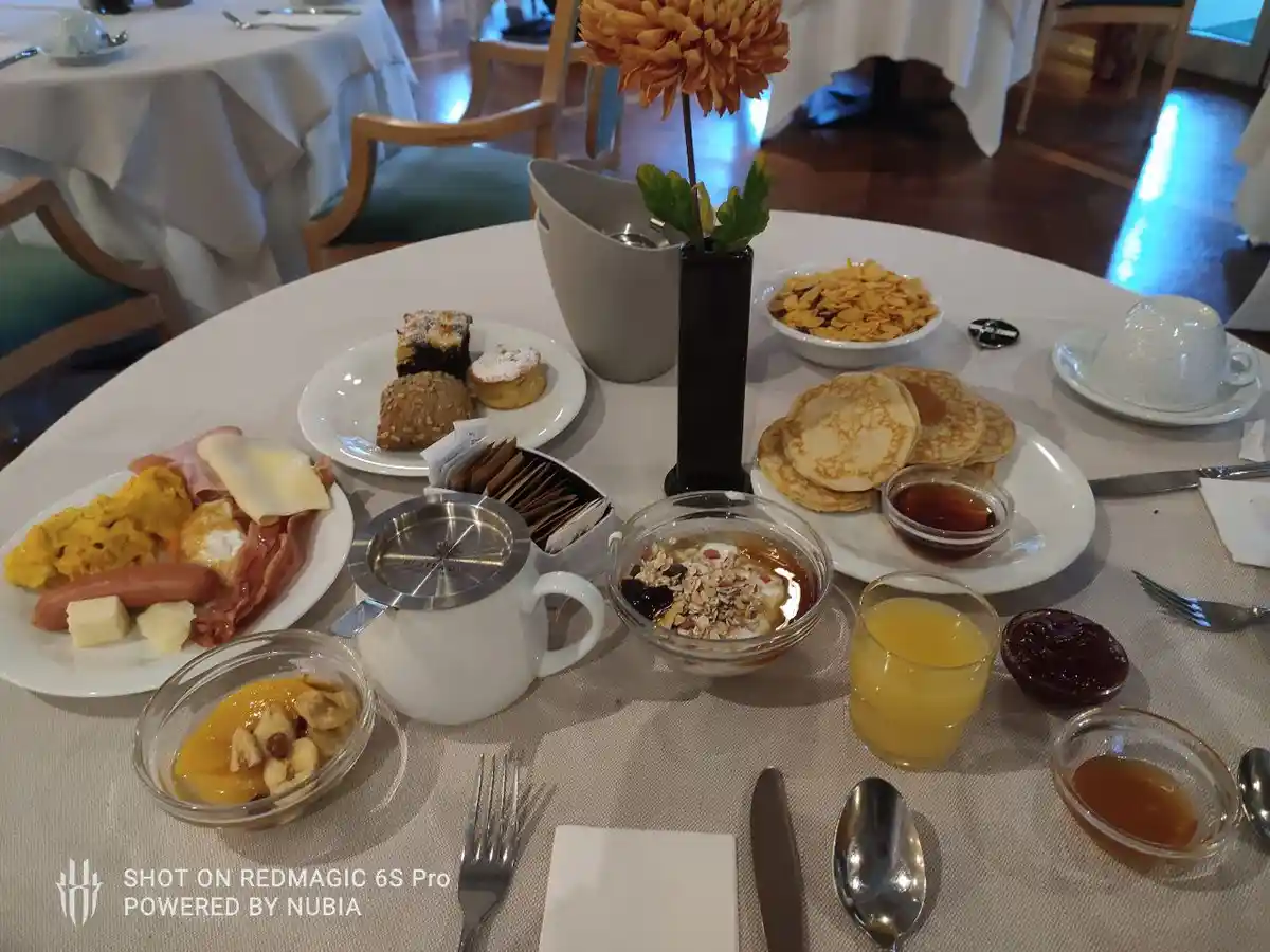 A lavish breakfast spread on a white tablecloth features pancakes, scrambled eggs, bacon, sausage, cereal, yogurt, orange…