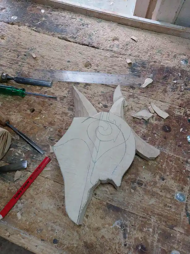 A partially carved wooden fox head sits on a cluttered workbench surrounded by wood shavings, chisels, a ruler, and a red…