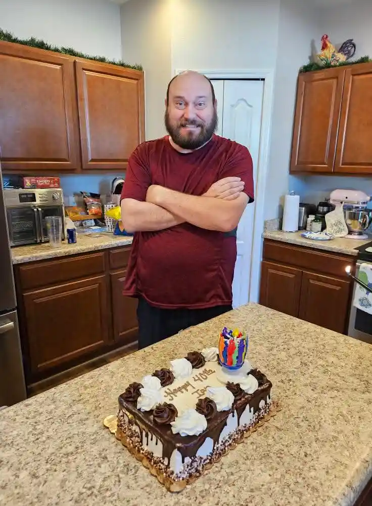 A bald man with a beard, wearing a red t-shirt and standing with arms crossed, smiles confidently in a kitchen behind a…