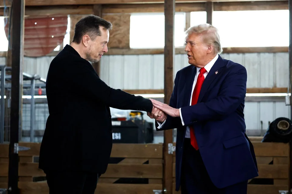Two men, Elon Musk and Donald Trump, are shaking hands in what appears to be a barn or industrial setting. Musk wears a…