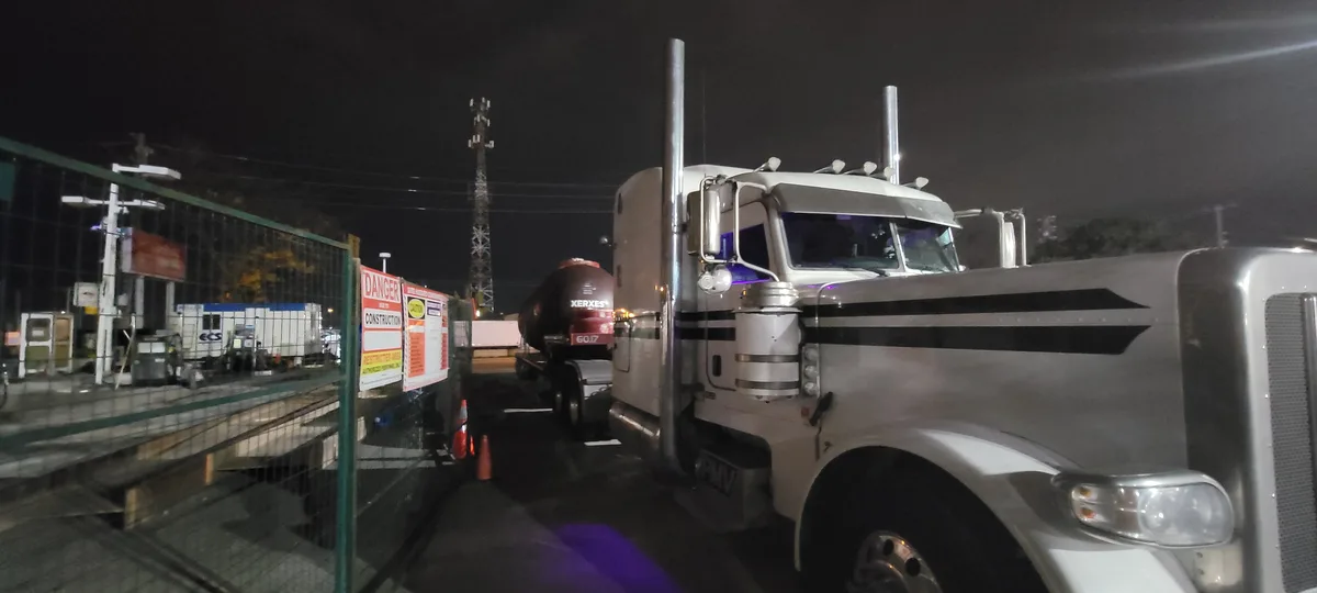 A large white semi-truck with chrome accents and purple underglow headlights is parked at night near a construction site…