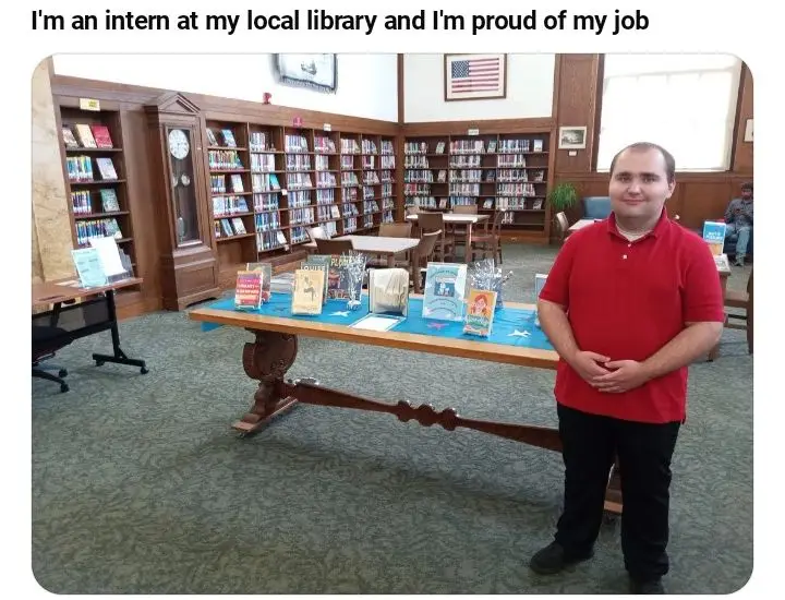 A man in a red polo shirt stands proudly in a library, smiling at the camera with his hands clasped. Behind him are tall…