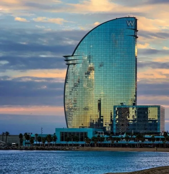 A tall, curved glass skyscraper with a prominent 'W' logo at its peak, reflecting the sky and surrounding buildings, stands…