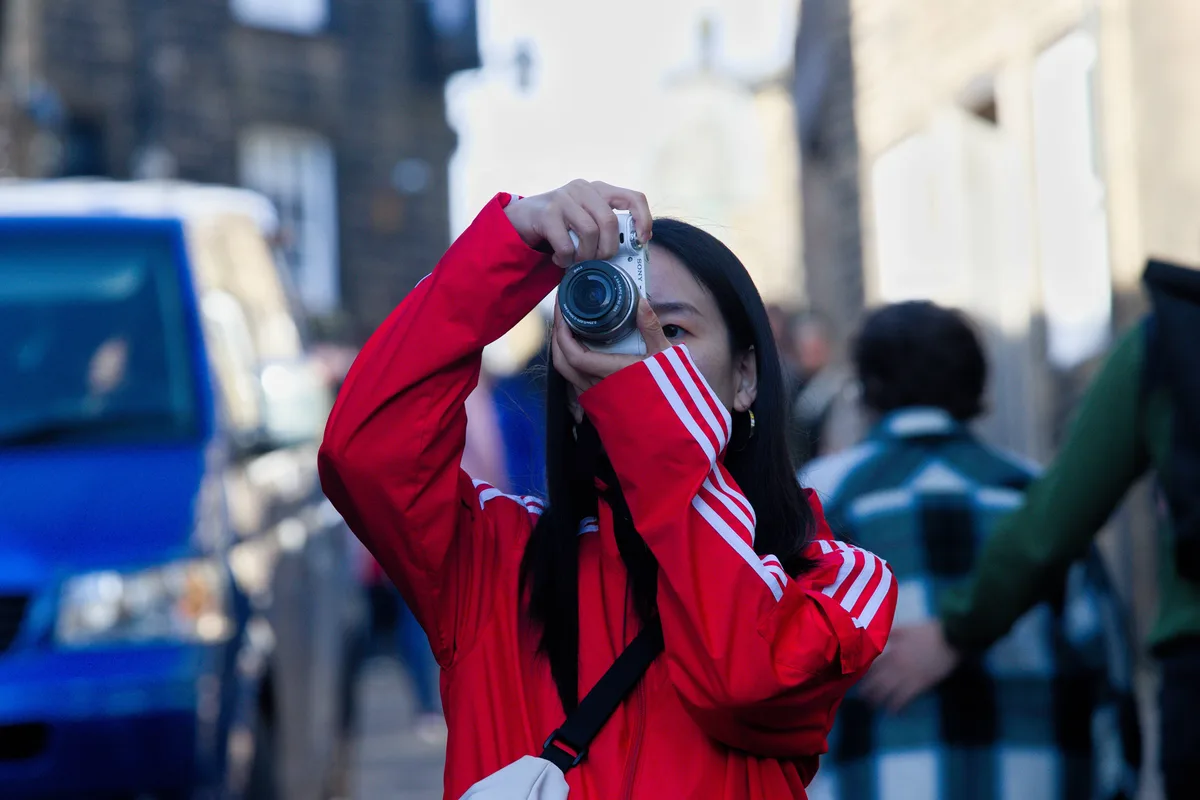 A person in a red jacket with white stripes holds a white Sony camera up to their face, partially obscuring their view as…