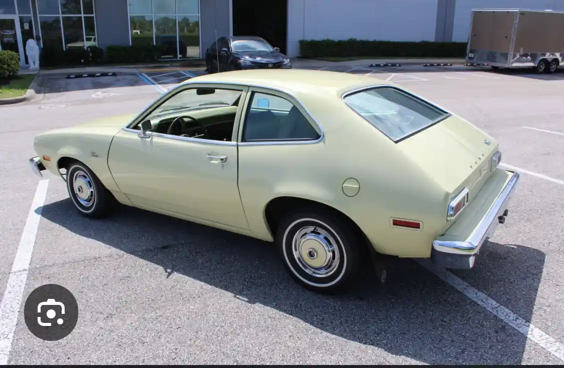 A pale yellow vintage Ford Pinto is parked in a lot, viewed from the rear three-quarter angle. The car has chrome bumpers…