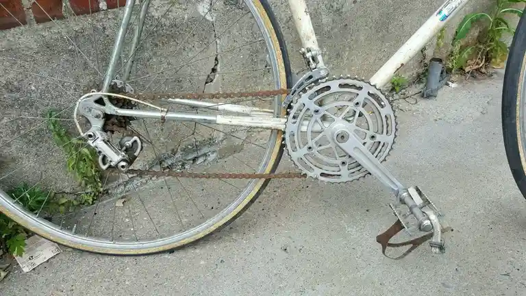 A close-up of a rusty, old bicycle's drivetrain, showing the chain, cassette, and crankset, with the bike leaning against a…