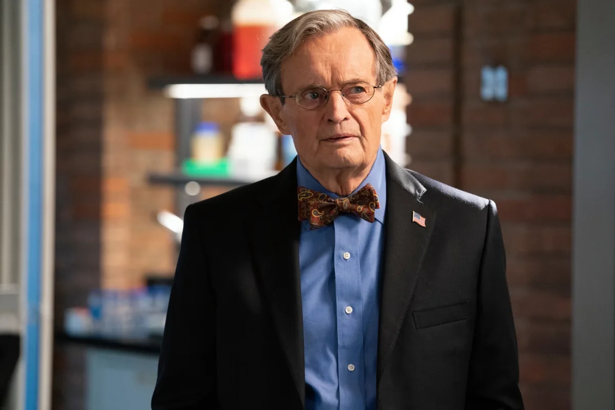 a man with glasses, a black suit, blue shirt, and paisley bow tie stands in a lab with a brick wall background. He has a…