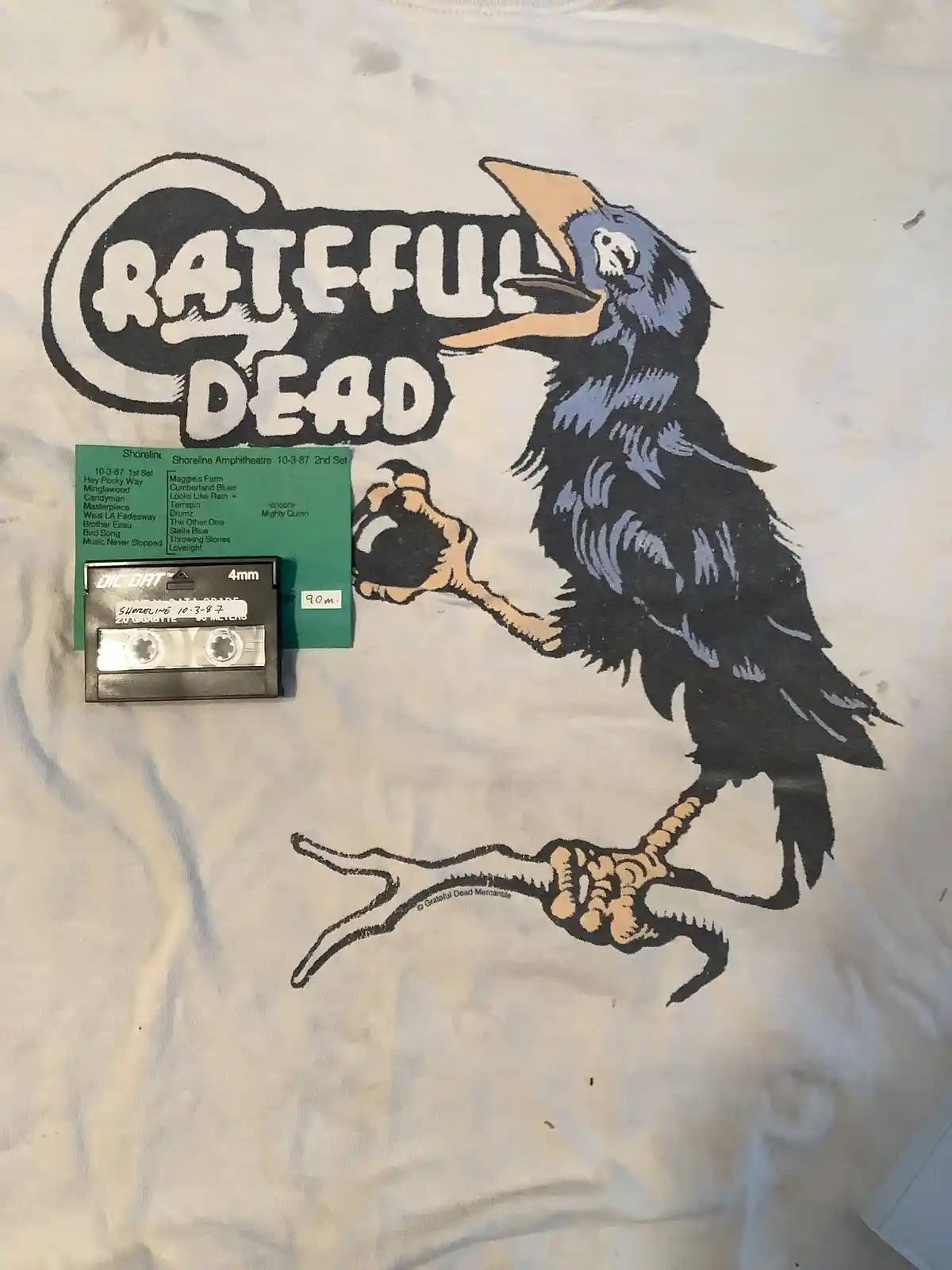 A white t-shirt with a Grateful Dead graphic of a crow holding a cassette tape, which is labeled 'Shoreline 10-3-87' and…