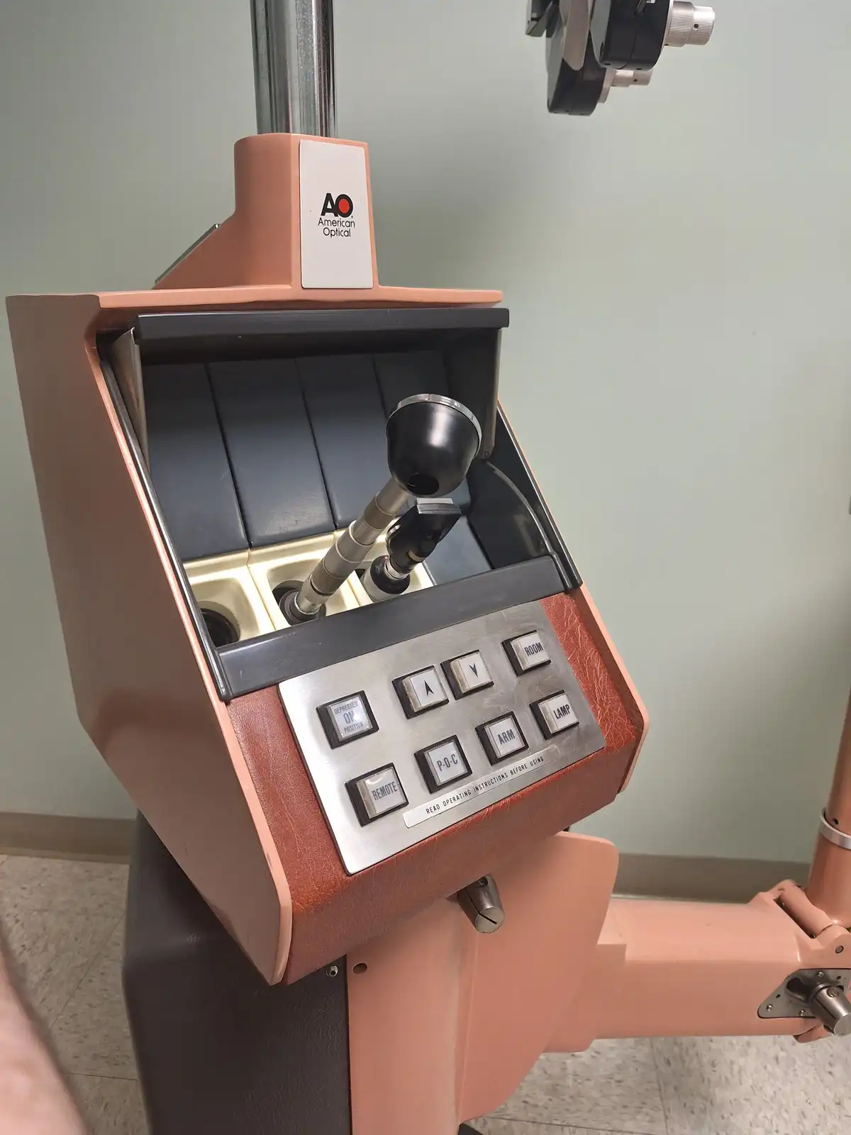 A close-up photo of an old-fashioned, peach-colored American Optical eye examination machine with a control panel featuring…