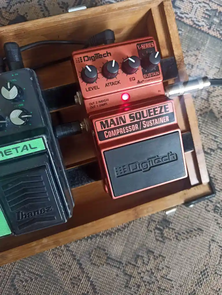 A close-up photo shows two guitar effects pedals in a wooden pedalboard. The prominent red Digitech 'Main Squeeze'…