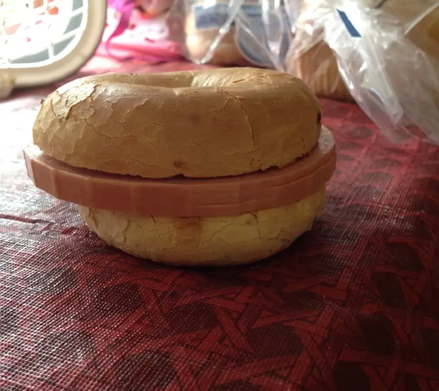 A close-up photo shows a bagel sandwich made with two bagel halves and a single slice of ham in between, sitting on a red…