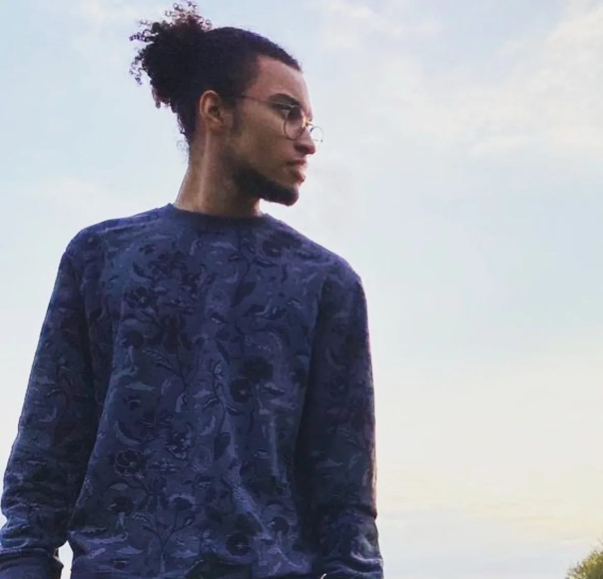 A man with curly hair in a bun, wearing round glasses and a dark blue patterned long-sleeve shirt, stands outdoors looking…
