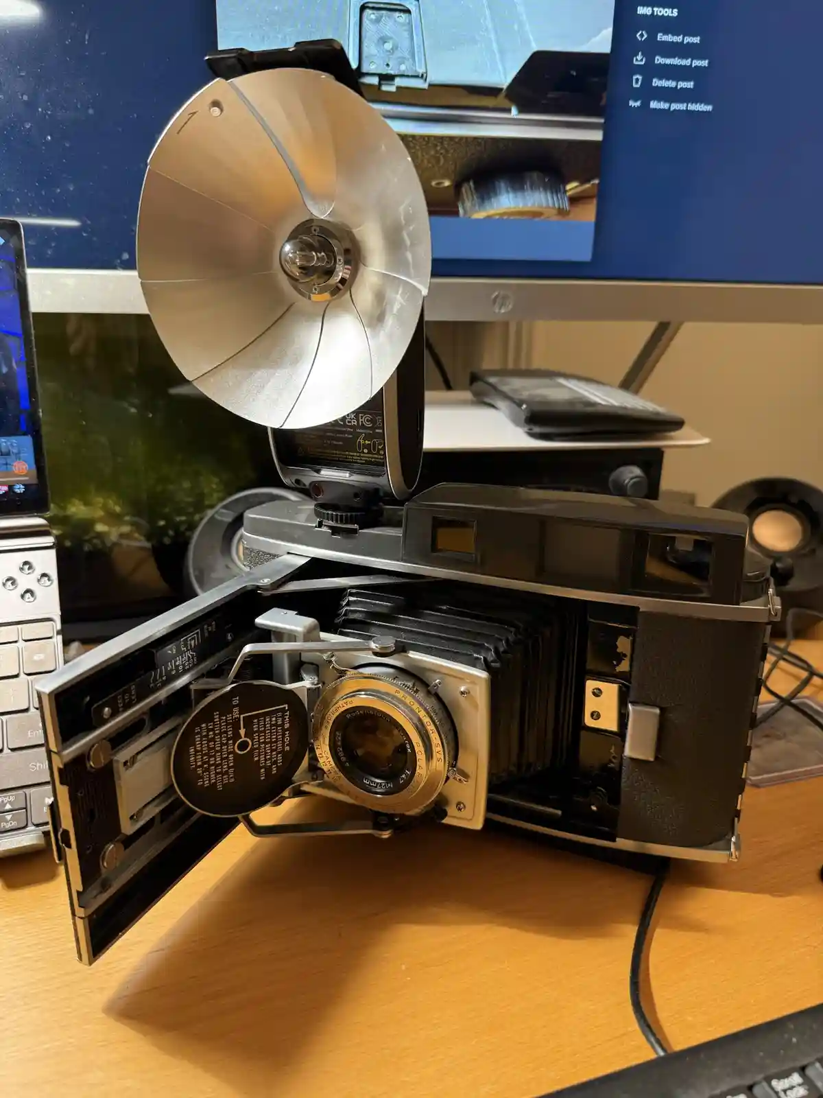 A vintage Polaroid camera with its back open, revealing the lens and internal mechanism, sits on a desk with a modern…