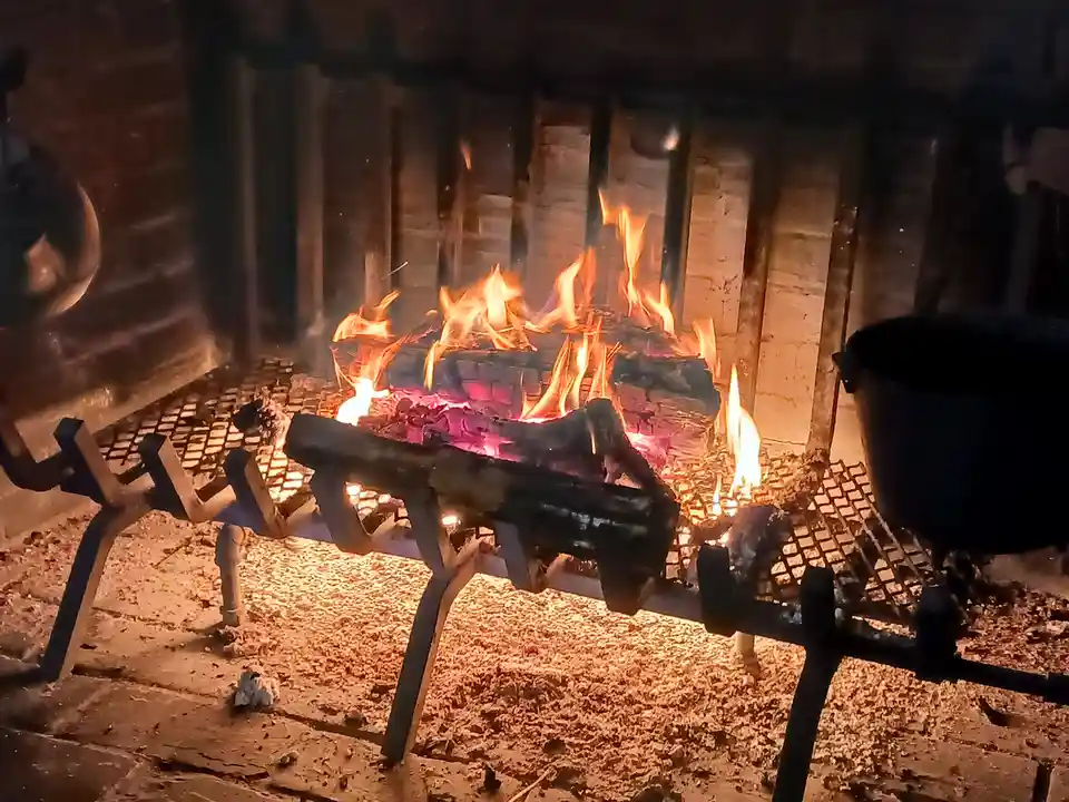 A close-up of a roaring fireplace with burning logs and glowing embers, viewed through a metal grate. A black pot sits on…