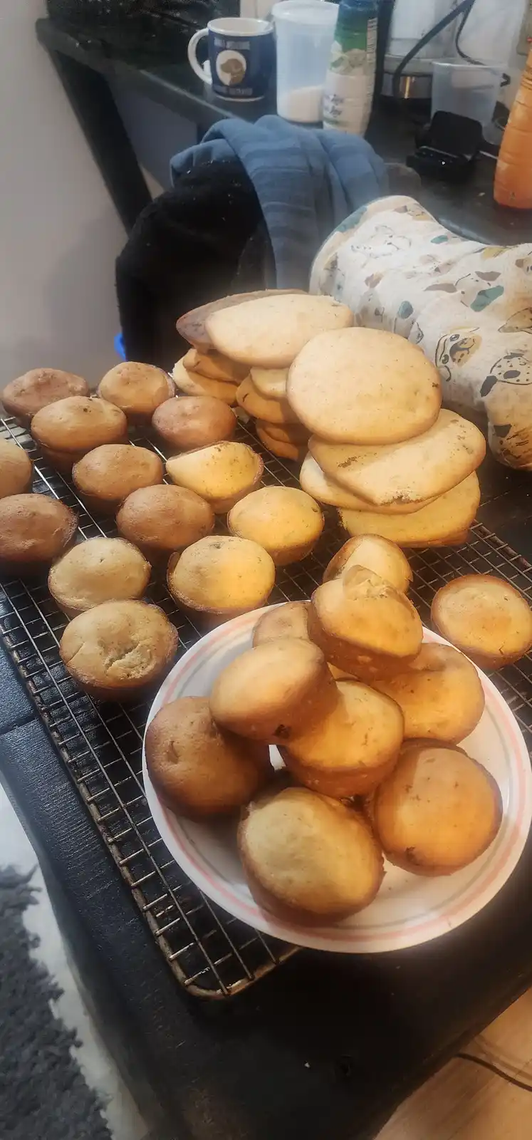 A kitchen counter displays numerous golden-brown muffins cooling on a wire rack and piled on a white plate with a pink rim…