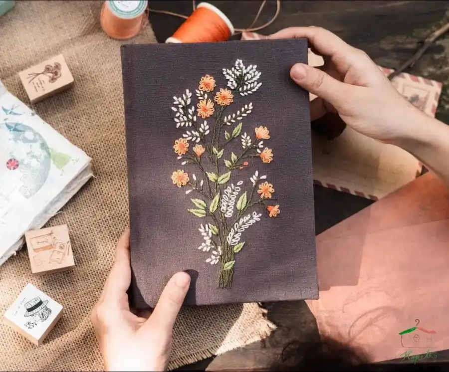 Two hands hold up a dark gray fabric-bound notebook with intricate floral embroidery in orange, white, and green…