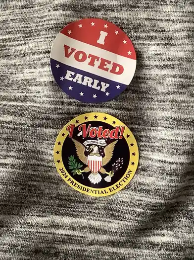 Two circular U.S. election-themed pins are pinned to a gray and white heathered fabric. The top pin is red, white, and blue…